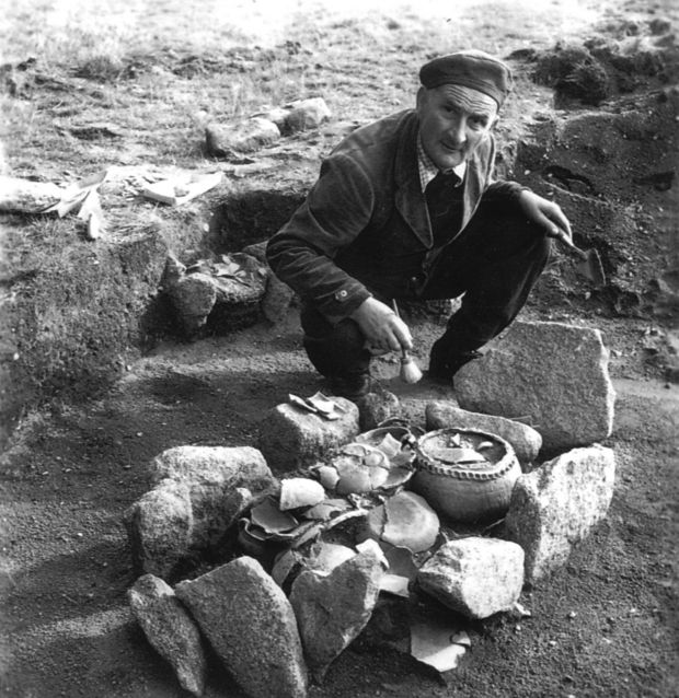 Schwarzweißfoto eines Mannes, der vor einem ausgegrabenen Grab sitzt. Das Grab besteht aus einer Steinfassung und ist mit vielen Keramikgefäßen gefüllt.